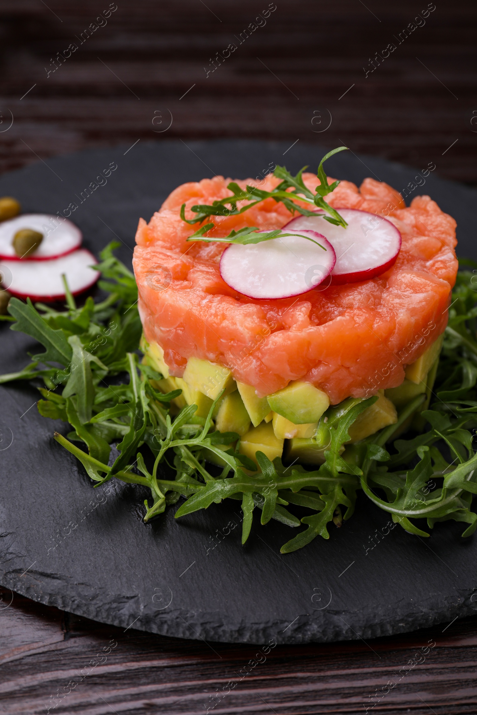 Photo of Tasty salmon tartare with radish, avocado and arugula on wooden table, closeup