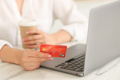 Photo of Online banking. Woman with credit card, paper cup and laptop paying purchase at white marble table, closeup