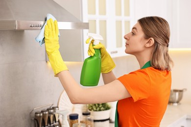 Photo of Professional janitor wearing uniform cleaning kitchen hood with rag and detergent indoors