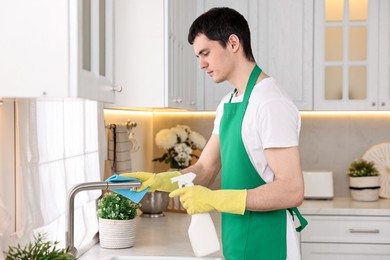 Photo of Professional janitor cleaning tap with rag and detergent in kitchen