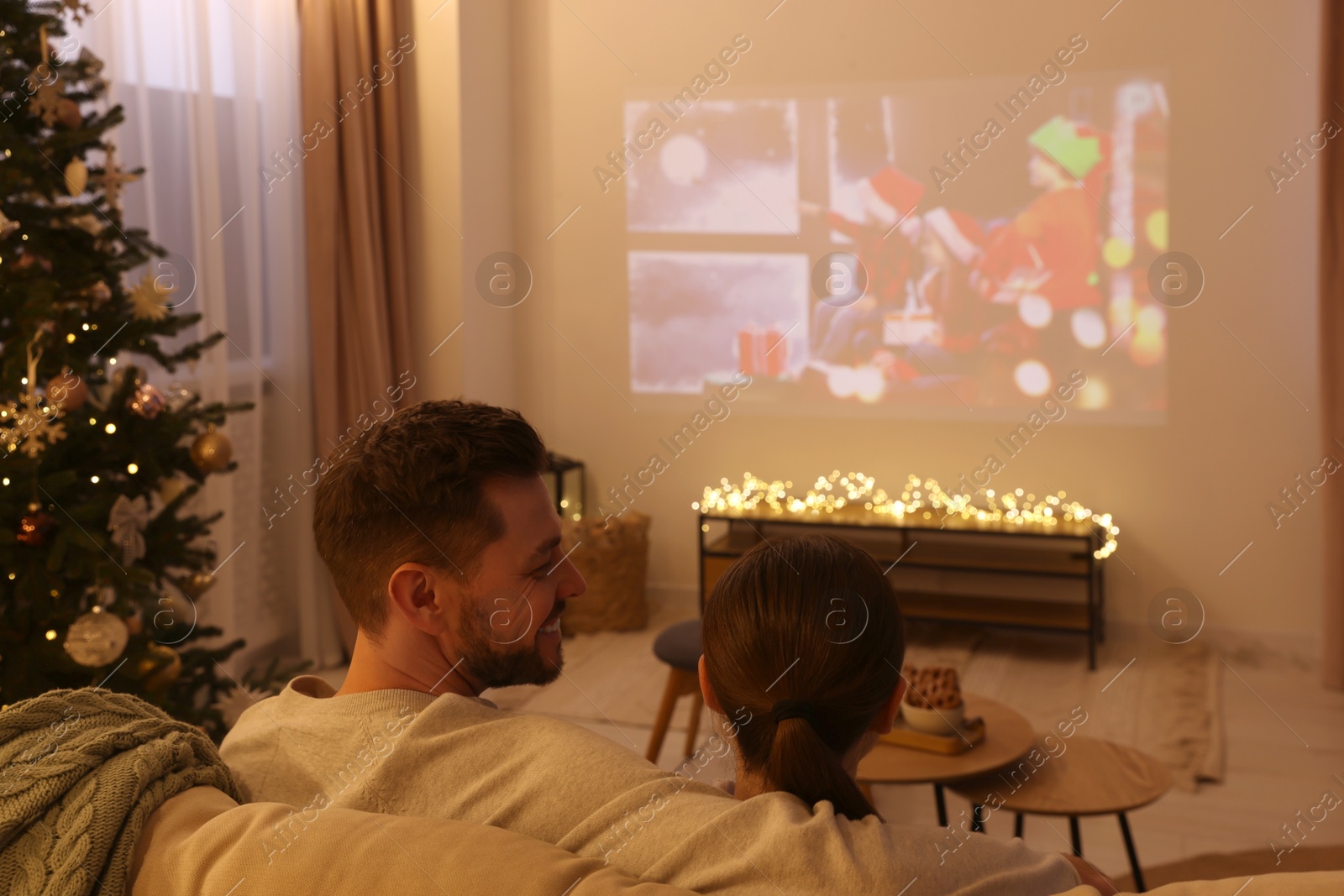 Photo of Couple watching Christmas movie via video projector in cosy room, back view. Winter holidays atmosphere