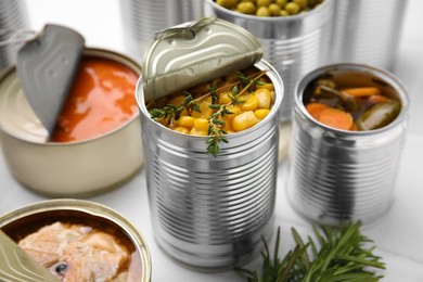 Open tin cans with corn kernels and different products on white table, closeup