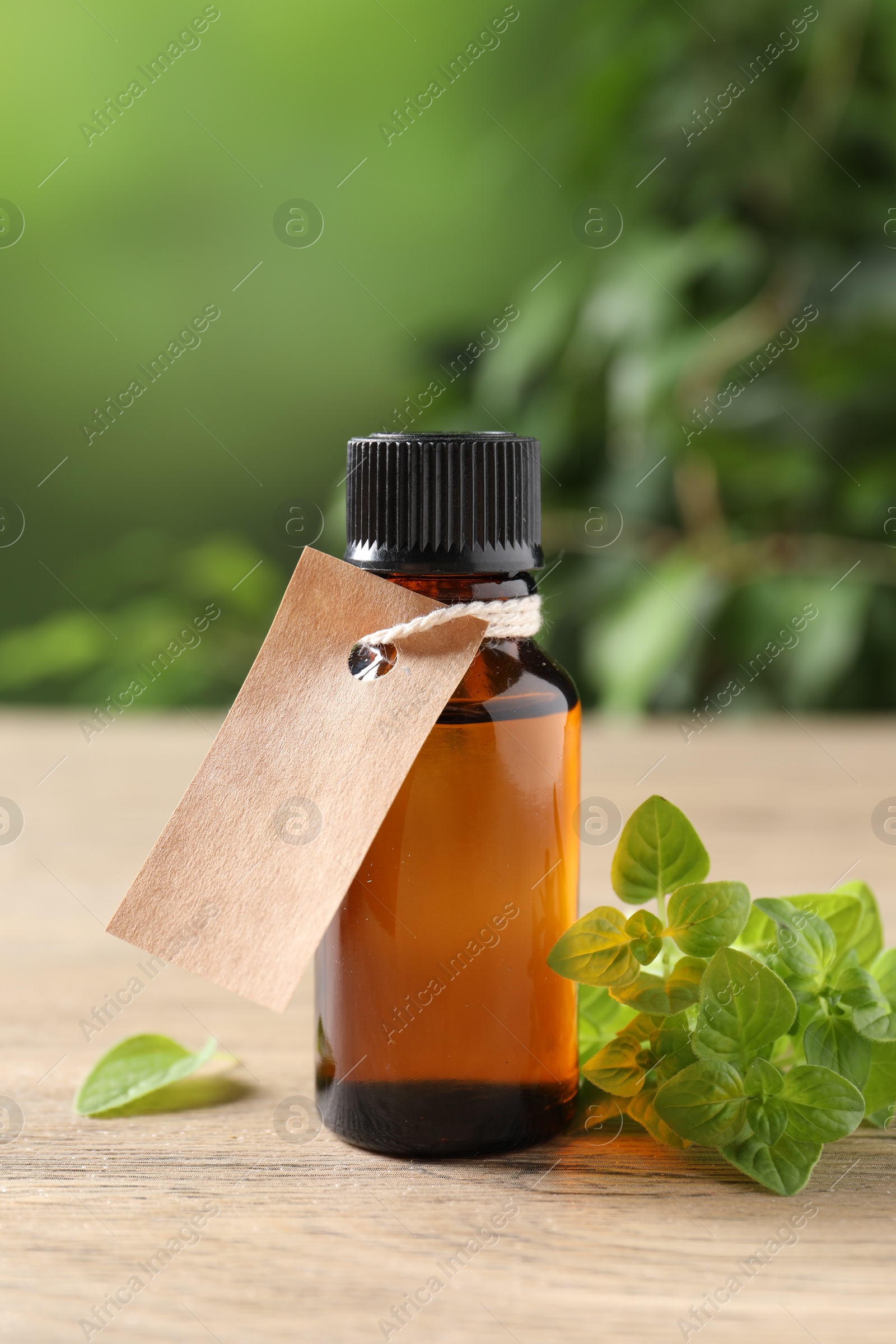 Photo of Essential oil in bottle with empty tag and oregano leaves on wooden table against blurred green background, closeup