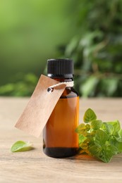 Essential oil in bottle with empty tag and oregano leaves on wooden table against blurred green background, closeup