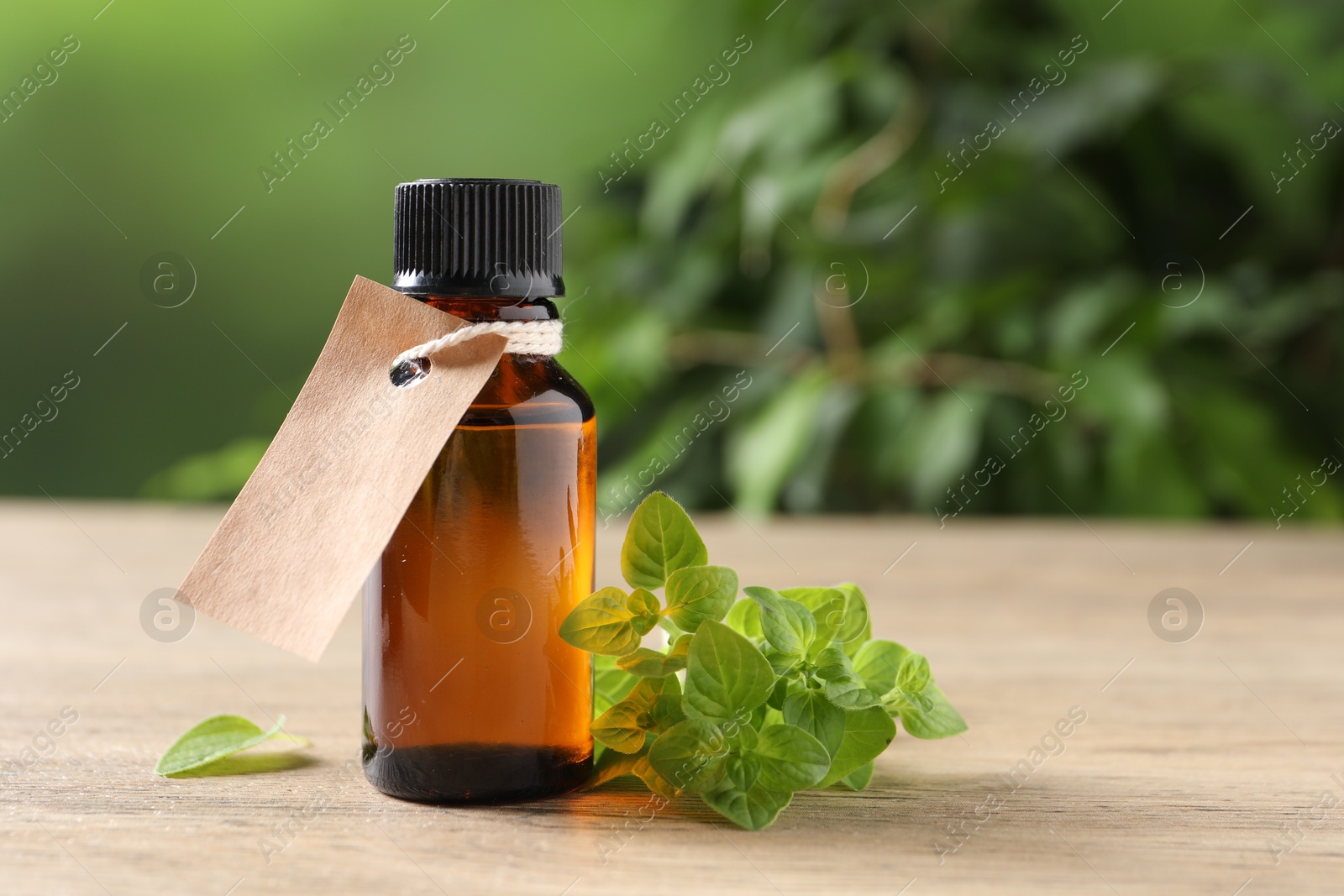 Photo of Essential oil in bottle with empty tag and oregano leaves on wooden table against blurred green background, closeup. Space for text