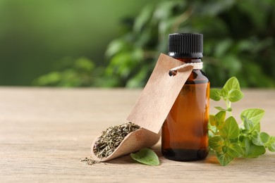 Essential oil in bottle with empty tag, dry herb and oregano leaves on wooden table against blurred green background, closeup. Space for text