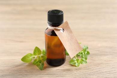 Essential oil in bottle with empty tag and oregano leaves on wooden table, closeup