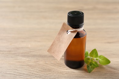 Essential oil in bottle with empty tag and oregano leaves on wooden table, closeup. Space for text