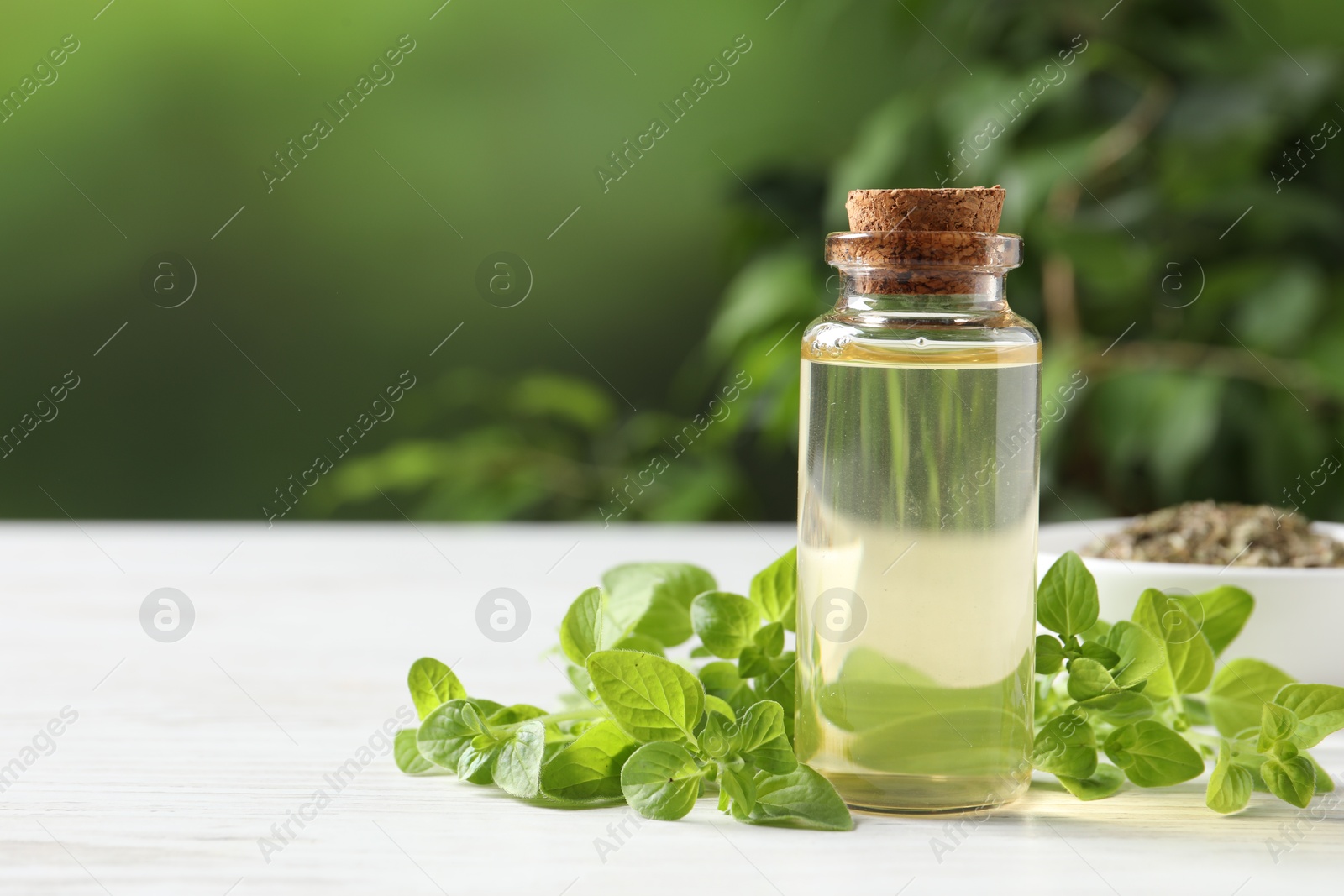 Photo of Essential oil in bottle and oregano leaves on white wooden table against blurred green background, closeup. Space for text