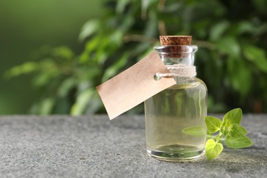 Essential oil in bottle with empty tag and oregano twig on grey textured table against blurred green background, closeup. Space for text