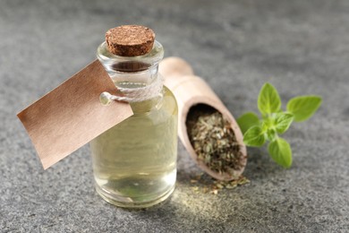 Essential oil in bottle with empty tag, dry herb and oregano twig on grey textured table, closeup