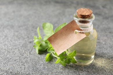 Photo of Essential oil in bottle with empty tag and oregano twigs on grey textured table, closeup. Space for text