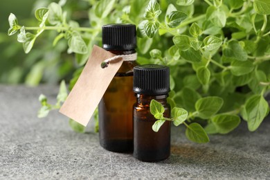 Essential oil in bottles, empty tag and oregano plant on grey textured table, closeup