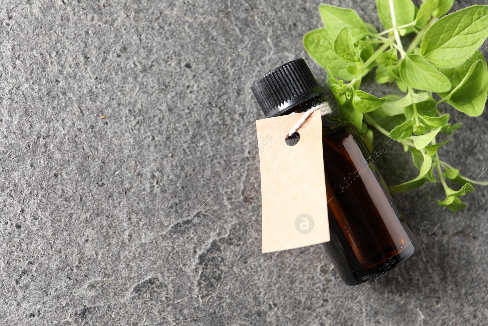 Photo of Essential oil in bottle with empty tag and oregano twig on grey textured table, flat lay. Space for text