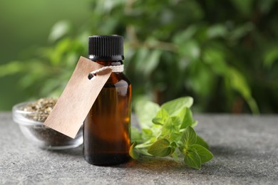 Photo of Essential oil in bottle with empty tag and oregano twig on grey textured table against blurred green background, closeup