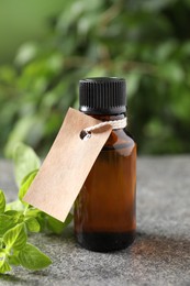 Essential oil in bottle with empty tag and oregano twig on grey textured table against blurred green background, closeup