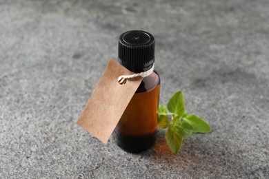 Essential oil in bottle with empty tag and oregano twig on grey textured table, closeup