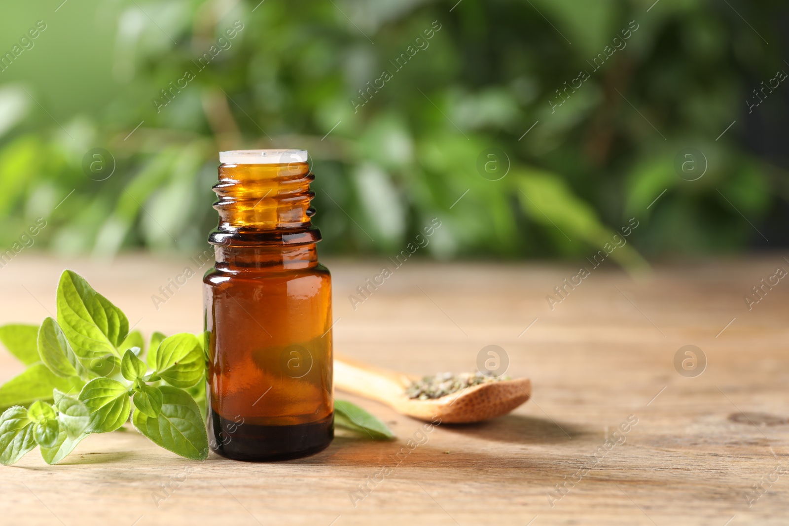 Photo of Essential oil in bottle, spoon with dry herb and oregano twigs on wooden table against blurred green background, closeup. Space for text