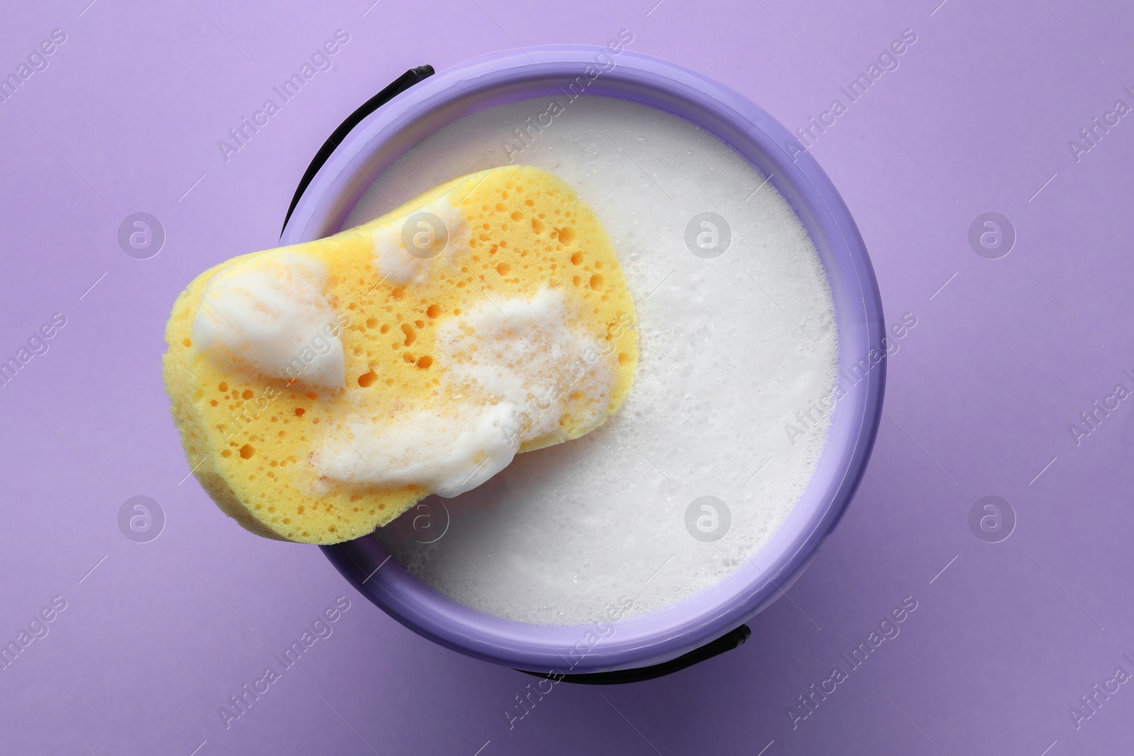 Photo of Bucket with foam and sponge on violet background, top view