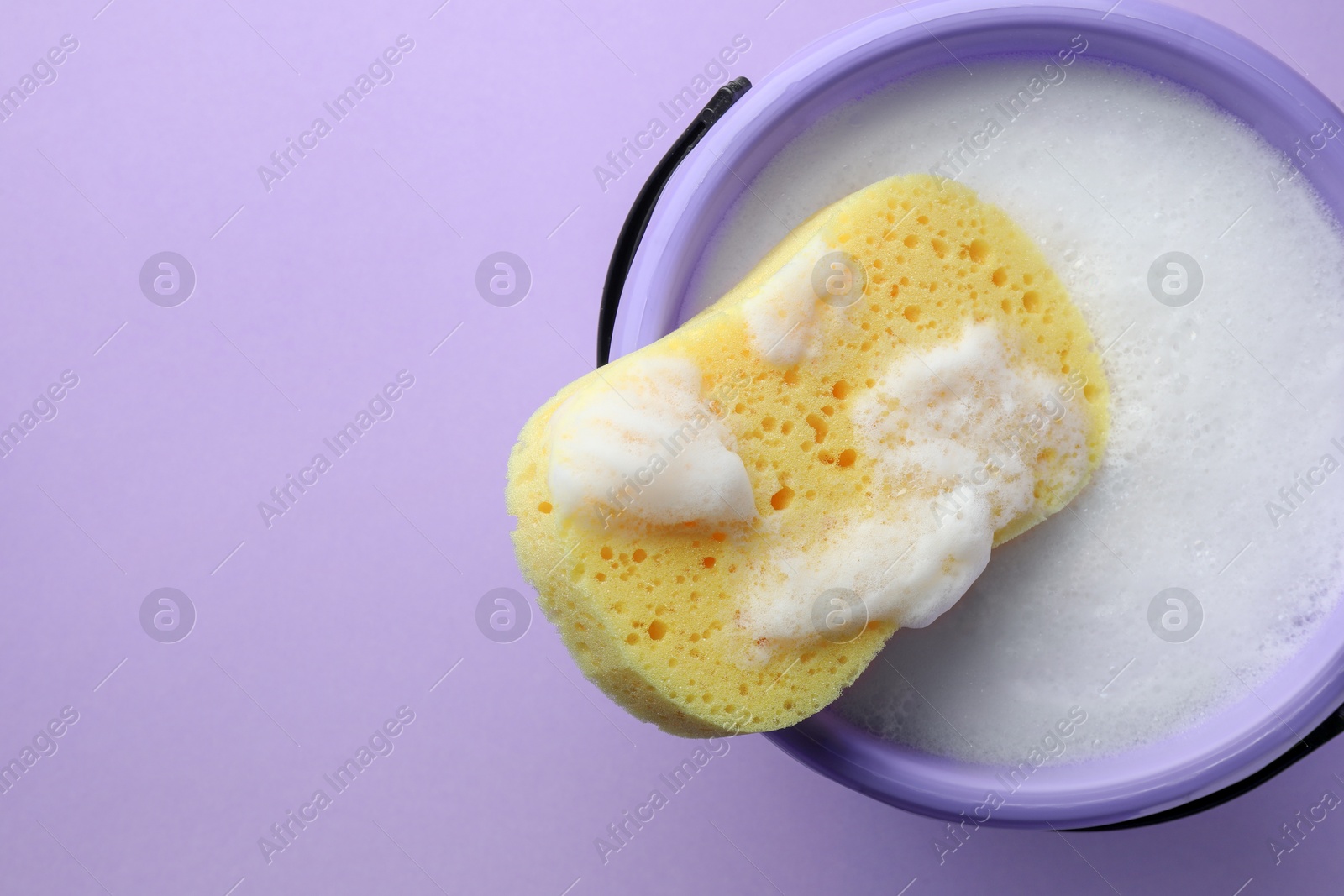 Photo of Bucket with foam and sponge on violet background, top view. Space for text