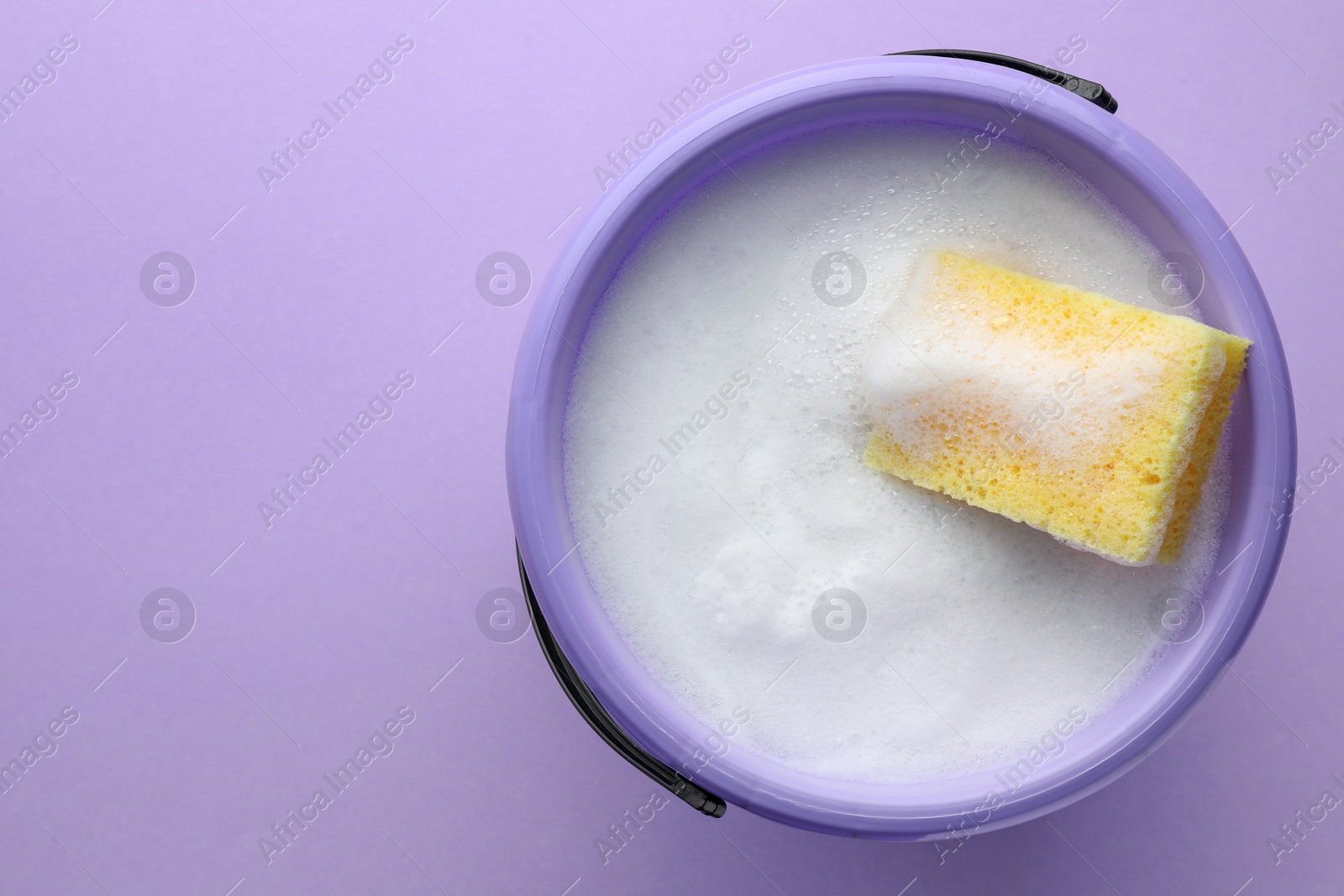 Photo of Bucket with foam and sponge on violet background, top view. Space for text