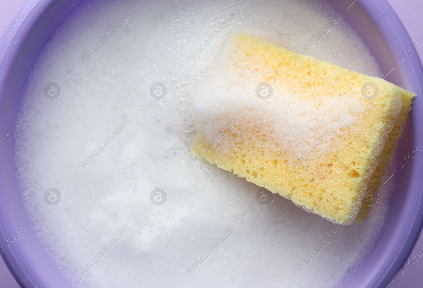 Photo of Bucket with foam and sponge on violet background, top view