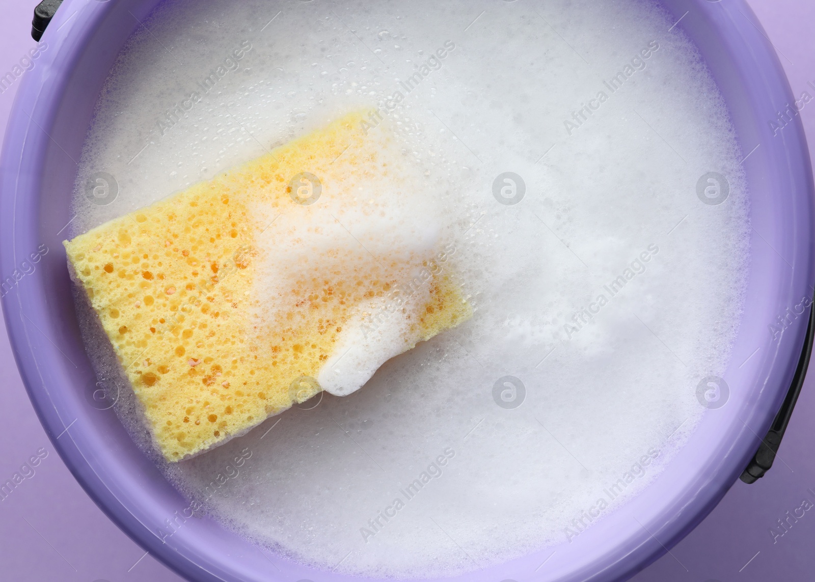 Photo of Bucket with foam and sponge on violet background, top view