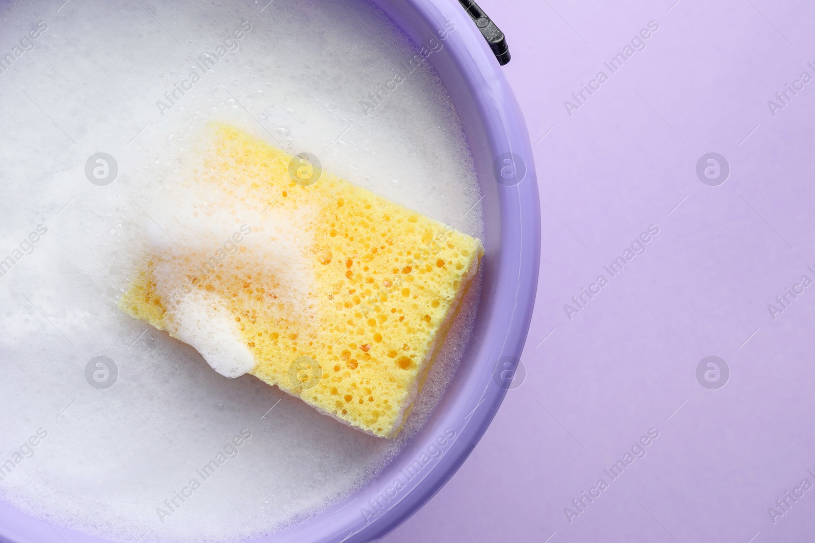 Photo of Bucket with foam and sponge on violet background, top view