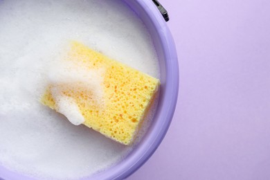 Photo of Bucket with foam and sponge on violet background, top view