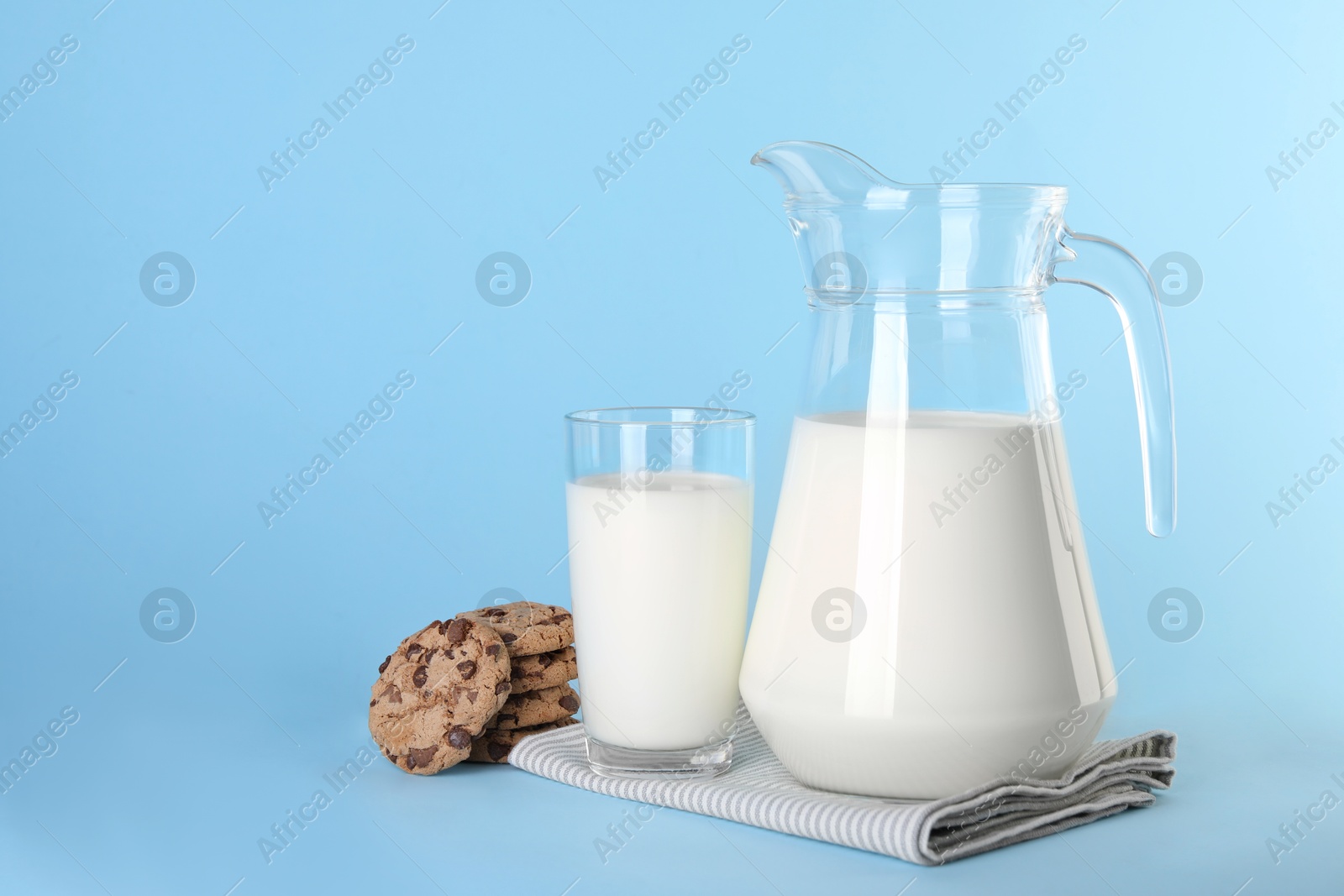 Photo of Jug of fresh milk, glass and cookies on light blue background, space for text