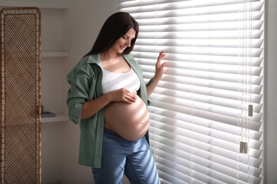 Beautiful pregnant woman near window blinds at home