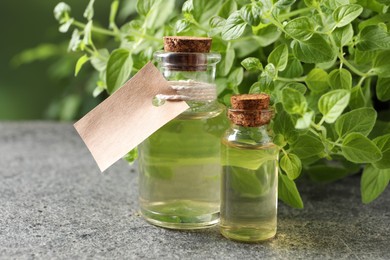 Essential oil in bottles, empty tag and oregano plant on grey textured table, closeup