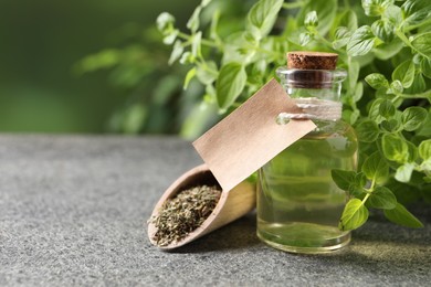 Photo of Essential oil in bottle with empty tag, dry herb and oregano plant on grey textured table against blurred green background, closeup. Space for text