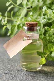 Essential oil in bottle with empty tag and oregano plant on grey textured table, closeup