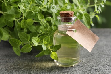 Essential oil in bottle with empty tag and oregano plant on grey textured table, closeup