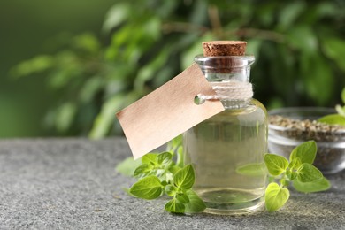 Photo of Essential oil in bottle with empty tag and oregano twigs on grey textured table against blurred green background, closeup. Space for text