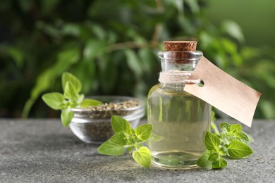 Essential oil in bottle with empty tag and oregano twigs on grey textured table against blurred green background, closeup