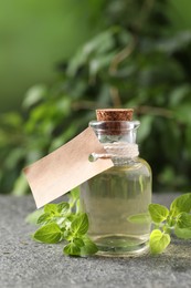 Essential oil in bottle, empty tag and oregano twigs on grey textured table against blurred green background, closeup