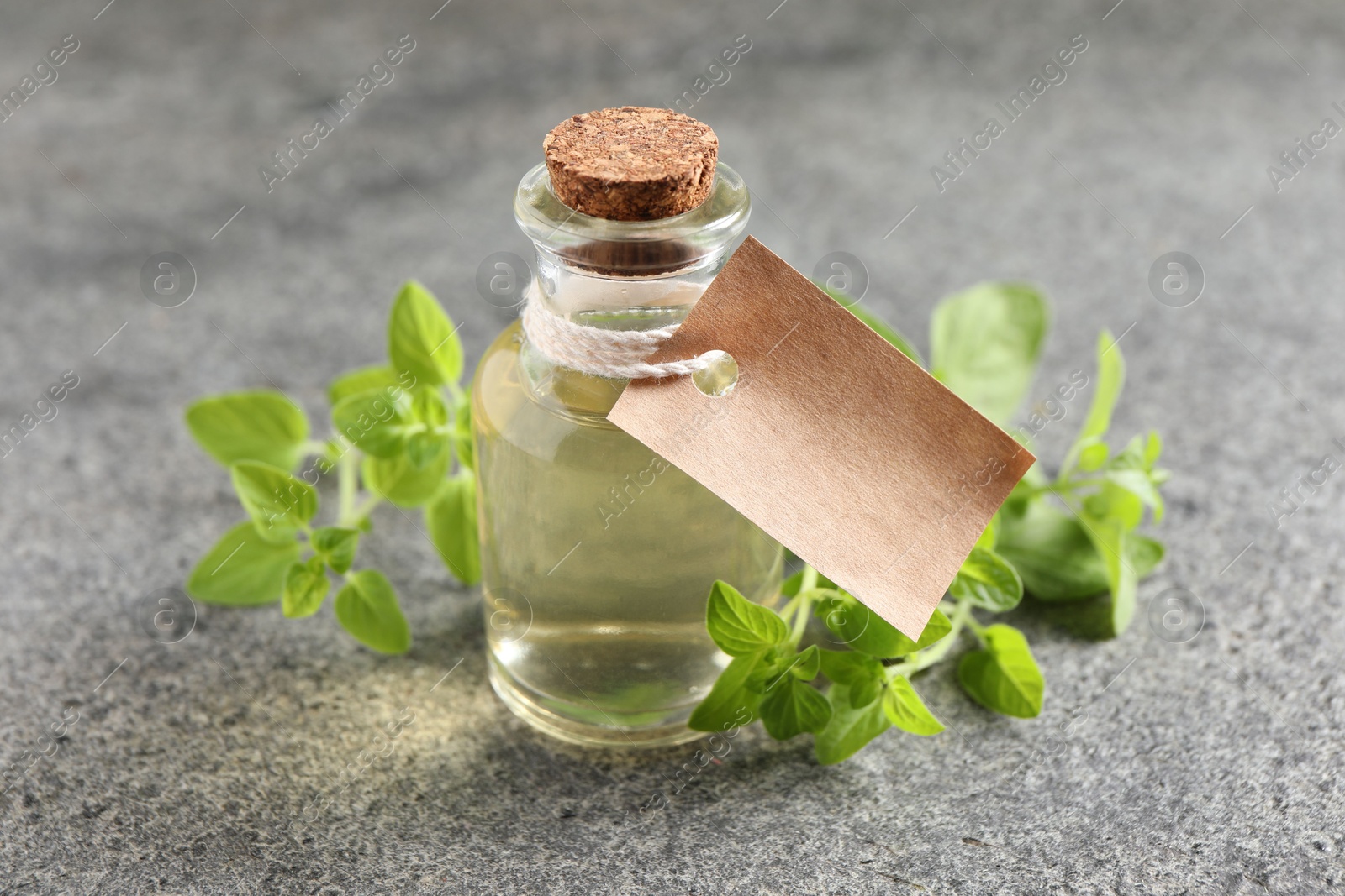 Photo of Essential oil in bottle with empty tag and oregano twigs on grey textured table, closeup