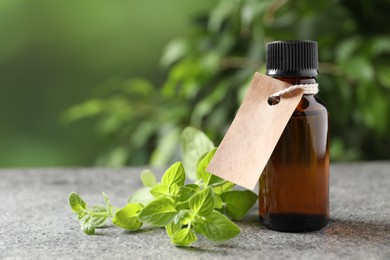 Essential oil in bottle with empty tag and oregano twig on grey textured table against blurred green background, closeup. Space for text