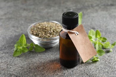 Essential oil in bottle with empty tag, dry herb and oregano twig on grey textured table, closeup