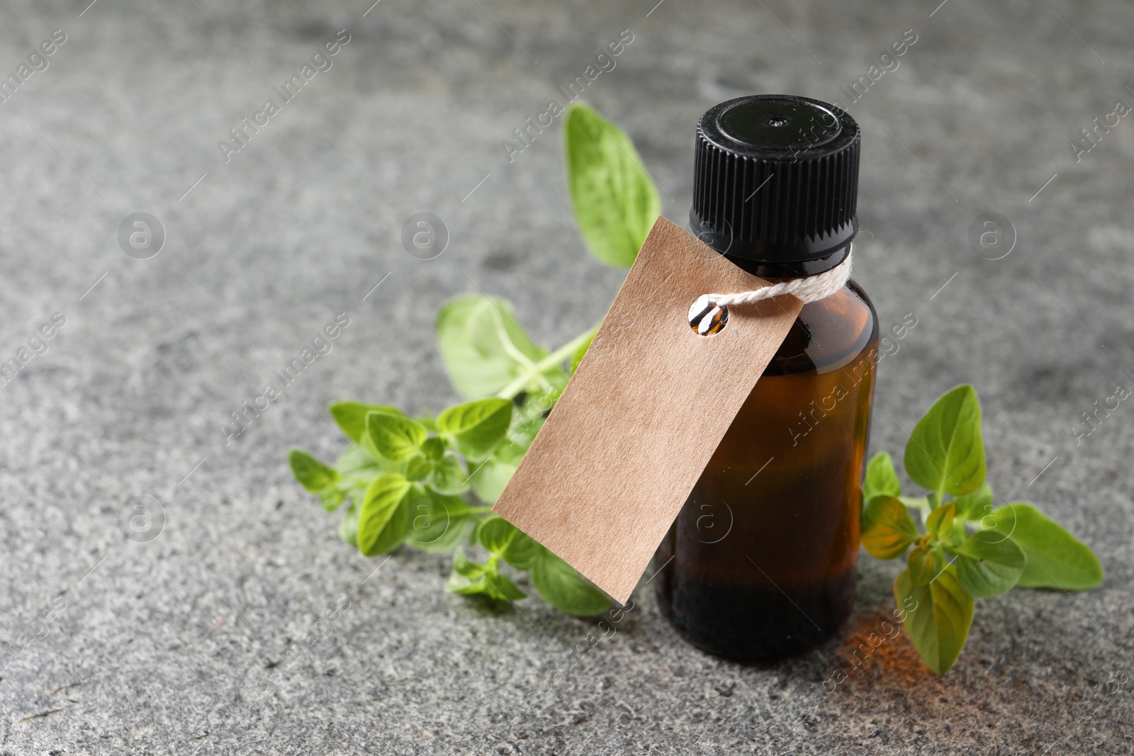 Photo of Essential oil in bottle with empty tag and oregano twig on grey textured table, closeup. Space for text