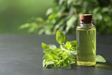 Photo of Essential oil in bottle and oregano twigs on dark textured table against blurred green background, closeup. Space for text