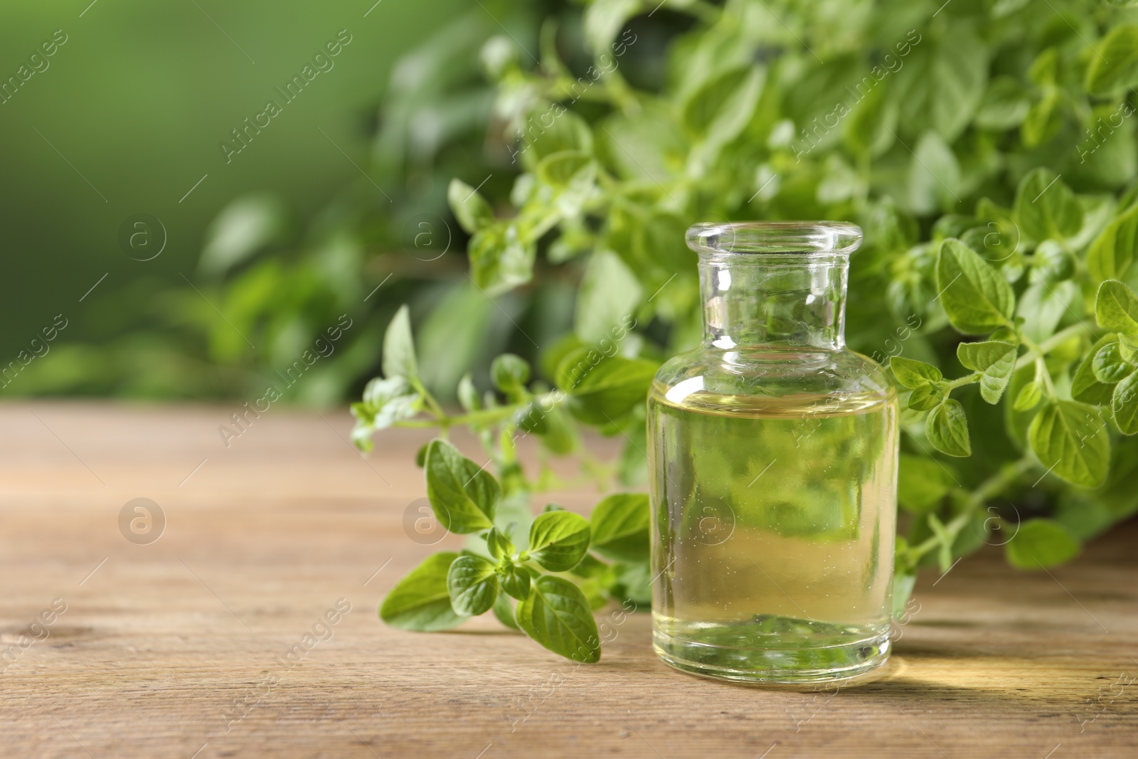 Photo of Essential oil in bottle and oregano twigs on wooden table against blurred green background, closeup. Space for text