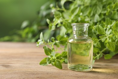 Essential oil in bottle and oregano twigs on wooden table against blurred green background, closeup. Space for text