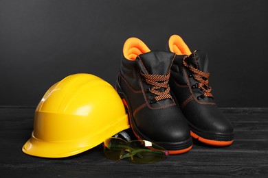 Photo of Pair of working boots, hard hat and goggles on black wooden surface