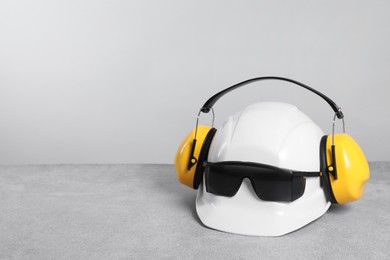 Photo of Hard hat with earmuffs and goggles on gray surface against light background, space for text