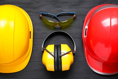Photo of Hard hats, earmuffs and goggles on black wooden background, flat lay