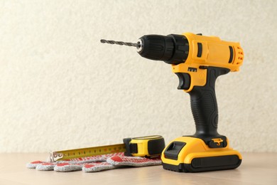 Cordless electric drill, gloves and measuring tape on wooden surface