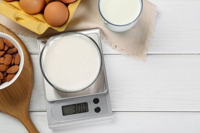 Photo of Kitchen scale with bowl of sugar among milk, almond and eggs on white wooden table, flat lay. Space for text